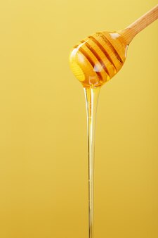 黄色​の​背景​に​木製​の​ハニーディッパー​から​金色​の​蜂蜜​が​滴り​落ちる​。​健康的​な​食生活​の​コンセプト​、​生態学的​に​純粋な​製品