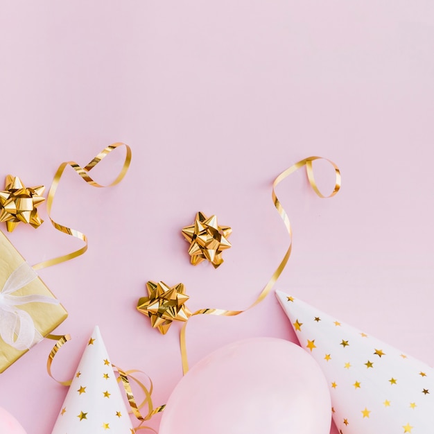 Golden ribbon bow; streamers; gift box; balloons and party hat on pink background