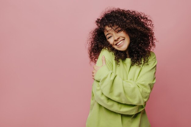 Good caucasian young woman with curly hair hugs herself with two hands on pink background Brunette smiles with teeth and wears light green sweatshirt Lifestyle different emotions leisure concept