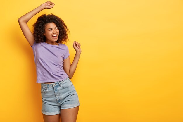 Good humored African American slim girl dances on yellow background, looks happily away, wears casual purple t shirt and jean shorts