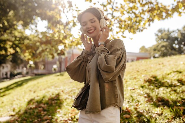 Good humored girl in white jacket with handbag listening to music outdoors Cool woman with short hair in headphones smiling outside