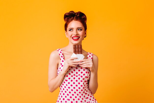 Добродушная европейская девушка ест шоколад. Молодая женщина кинозвезды в платье в горошек, улыбаясь на желтом пространстве.