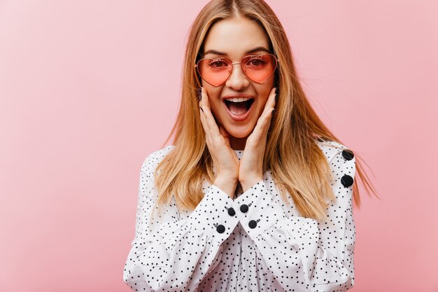 Good-humoured female model in sunglasses posing with surprised smile. Indoor portrait of glad trendy woman laughing on pink