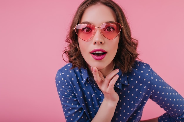 Good-humoured female model with brown curly hair posing in funny sunglasses. Indoor photo of pleased white girl standing on pink wall with interested face expression.