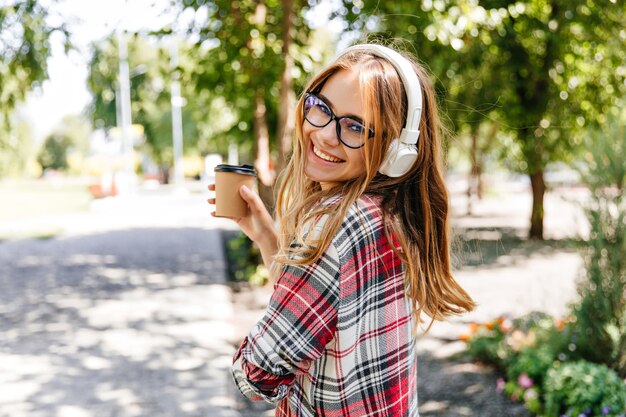 Good-humoured girl drinking coffee in park. Laughing blonde woman in glasses listening music on nature.
