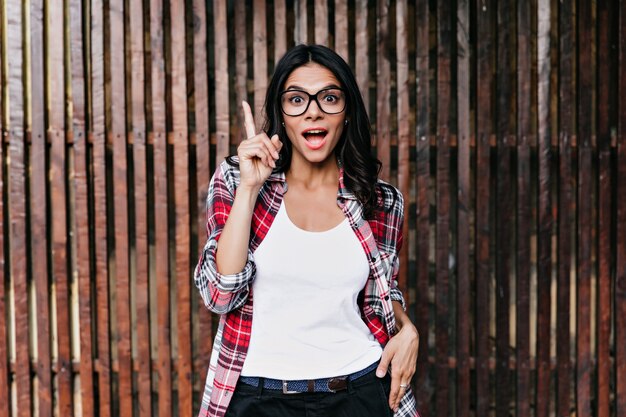 Good-humoured girl has a good idea. Outdoor photo of emotional latin lady isolated on wooden wall.