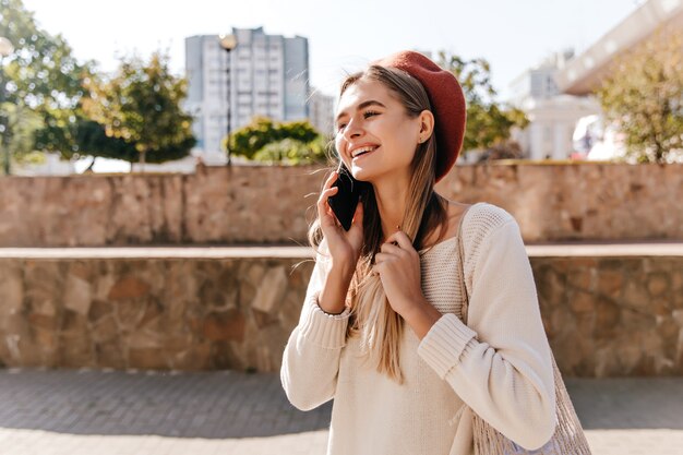 Добродушная девушка с длинными волосами разговаривает по телефону на улице. Привлекательная белая женщина в берете с удовольствием на открытом воздухе.