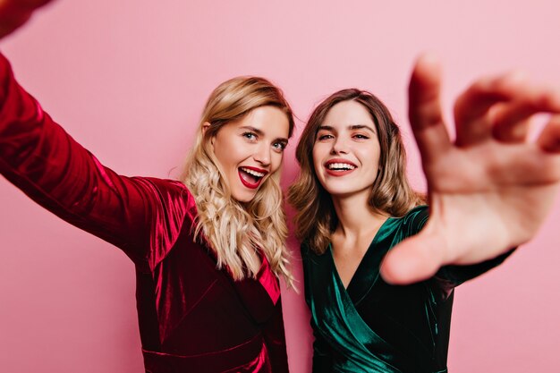 Good-humoured lady in red outfit fooling around with best friend. Glamorous caucasian girls preparing for party.