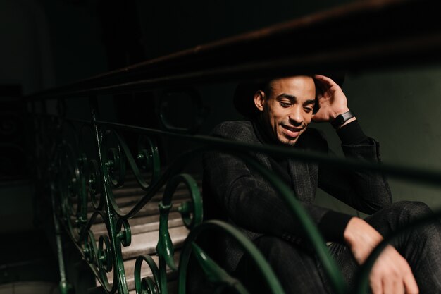 Good-humoured well-dressed man sitting on steps. Portrait of handsome african guy posing with pensive smile.