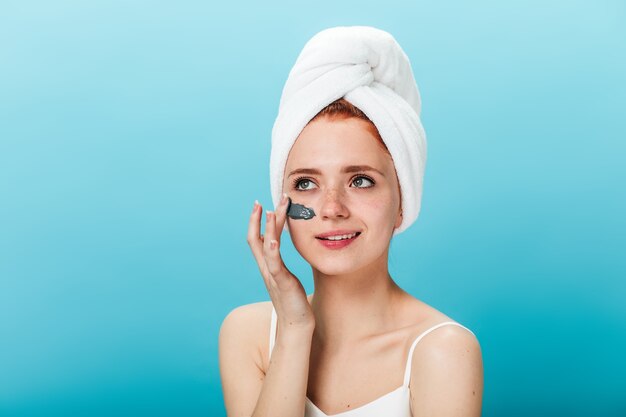 Good-humoured woman applying face mask. Studio shot of cheerful girl with towel on head doing spa treatment.