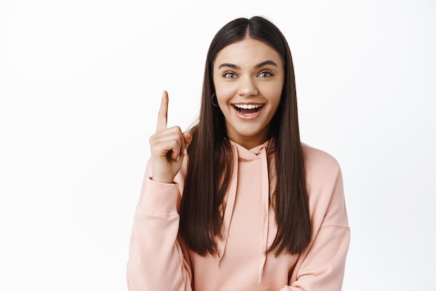 Good idea Excited brunette girl have a plan raising finger up and smiling suggesting interesting solution standing in hoodie against white background