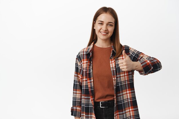 Good job. Young smiling woman happy about result, thumbs up in approval, like and agree, support your choice, standing satisfied against white background