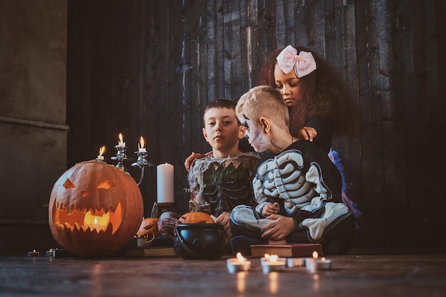 Good little friends in Halloween costumes are sitting on the floor with pumpkins and candles.