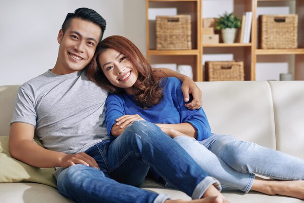 Красивая азиатская пара отдыхает вместе на диване у себя дома и улыбается