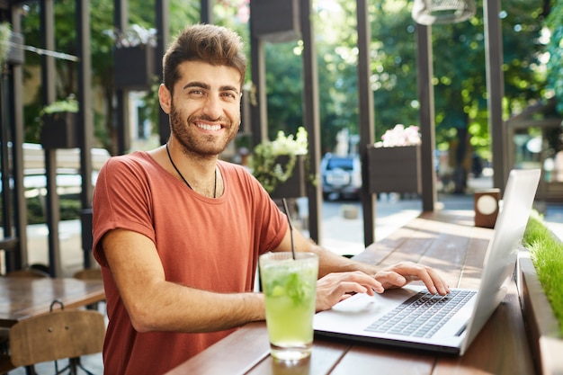 Good-looking, attractive bearded man broadly smiling at camera dressed casually sitting at wooden table drinking lemonade, surfing high-speed internet on laptop pc. Enjoying summer day.