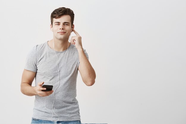 Привлекательный молодой человек в серой футболке с приятным видом держит мобильный телефон в руке, надев белые наушники, с радостью слушает любимые песни, используя музыкальное приложение.
