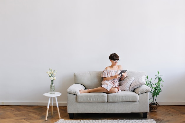 Good-looking barefooted girl in glasses lies on sofa with new gadget enjoying weekend