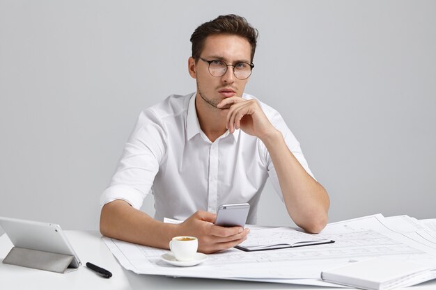 Good looking bearded male contractor sitting at desk in modern office interior using mobile phone while studying blueprint, having thoughtful serious look, touching chin. People, job and occupation