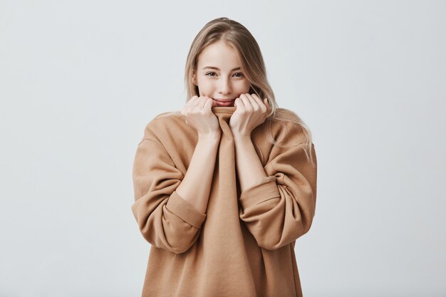 Good-looking beautiful woman smiling, pleased to hear compliments, isolated. Pretty female with fair hair and dark appealing eyes hiding face in sweater, being shy