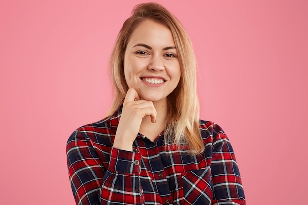 Good looking blonde female with gentle smile, keeps one hand under chin, dressed in checkered shirt