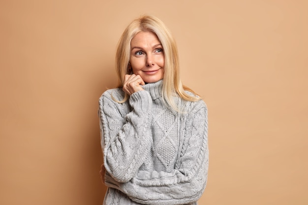 Good looking blonde mature European woman stands in thoughtful pose wears comfortable grey jumper concentrated aside ponders on making choice.