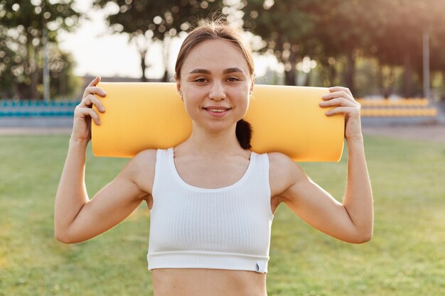 Good looking brunette female wearing white to holding yellow karemat on shoulders, looking smiling directly at camera, training outdoor in stadium in sunny day.