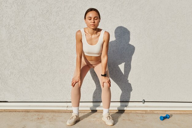 Good looking brunette female of young age wearing white top and beige leggins, standing with closed eyes, breathing after sport exercises or running, health care.