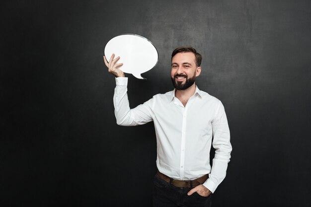 Good-looking brunette man holding blank speech bubble and looking at camera over dark gray wall copy space