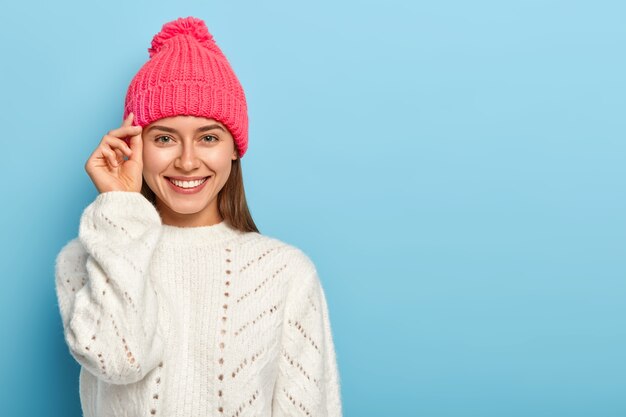 Good looking brunette young woman keeps hand near face, smiles pleasantly, wears hat and white knitted sweater,  expresses good emotions, poses against blue studio wall