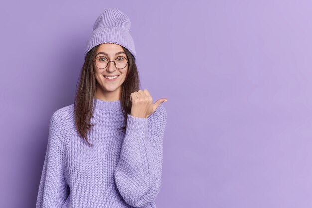 Good looking brunette young woman looks glad points thumb right wears purple hat knitted sweater and round glasses, pointing at copy space