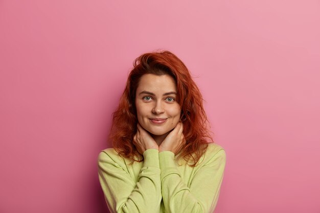 good looking calm woman with natural beauty, ginger hair, looks with satisfaction, keeps hands under face, dressed in casual jumper