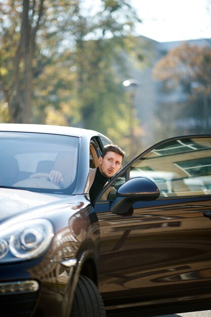 見栄えの良い白人ドライバーは、美しい晴れた日に街で開かれた車のドアから見ています。