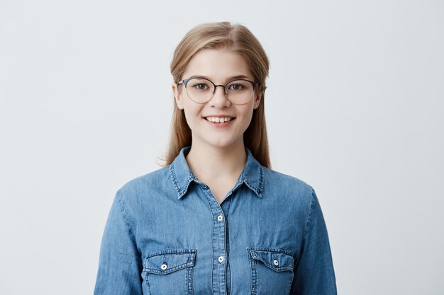 Good looking caucasian female with blonde straight hair, wearing glasses and denim shirt, smiles happily, has good mood after successful day at university, glad and pleased to pose 