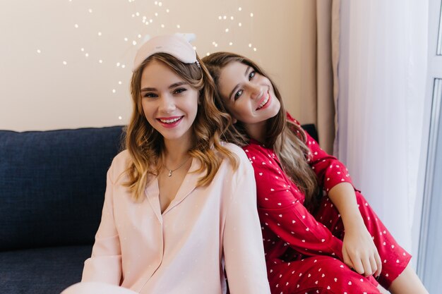 Good-looking caucasian ladies spending morning on sofa. Indoor portrait of smiling female friends in pajamas posing on cozy couch.