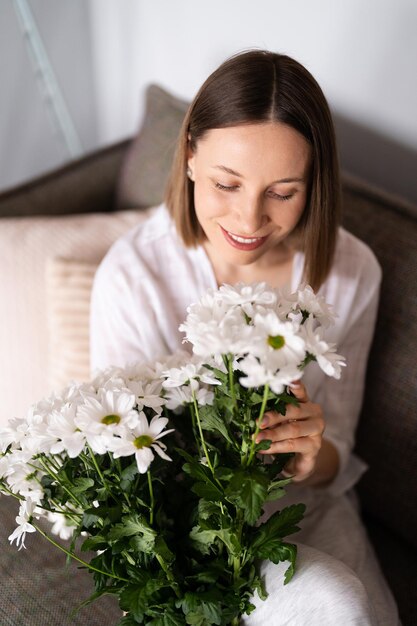 잘 생긴 백인 여성은 소파에서 휴식을 취하면서 신선한 흰 국화 꽃다발을 받는 것을 기쁘게 생각합니다.