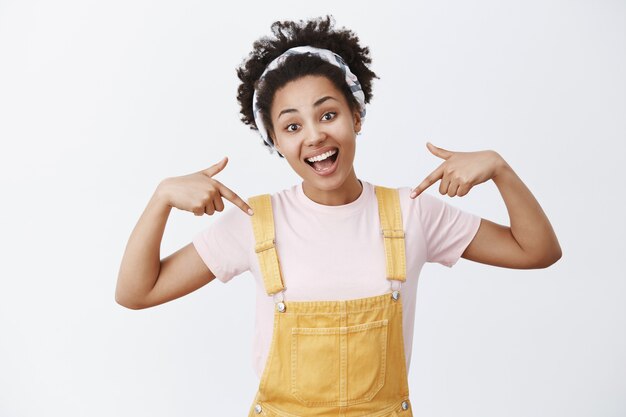 Good-looking charming active and optimistic African American girl in yeloow overalls and headband pointing at herself, tilting head and smiling broadly, being proud of her deeds over grey wall