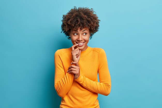 Good looking cheerful Afro American woman smiles gently wears casual long sleeved jumper looks joyful aside poses against blue wall