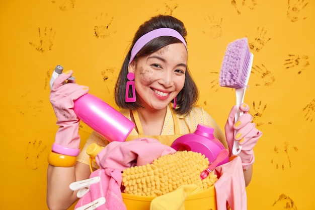 Good looking cheerful asian woman smiles happily holds brush and spray detergent of cleaning liquid busy doing laundry wears protective rubber gloves has dirty face and clothes poses indoor