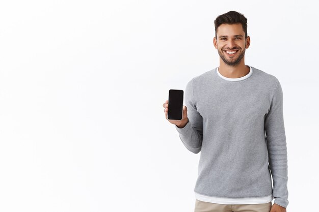 Красивый веселый молодой человек продвигает приложение для смартфона, держит телефон или что-то на экране мобильного, удовлетворенно улыбаясь