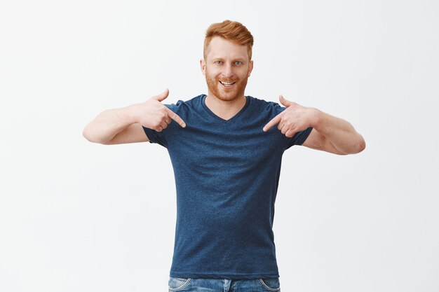 Good-looking confident and proud redhead male model with bristle in blue t-shirt, pointing at himself with index fingers and smiling