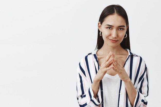 Good-looking confident and tricky woman in striped blouse, holding fingers together and smirking, having some bad intention in mind, planning to commit crime