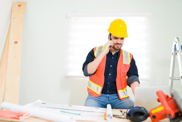 Good-looking contractor talking on the phone with a customer while typing on a laptop. Hispanic engineer solving construction problems