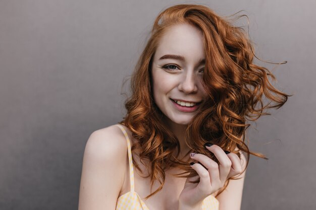 Симпатичная кудрявая девушка с большими глазами играет с рыжими волосами. Фотография потрясающей рыжеволосой дамы изолированной на серой стене с улыбкой.