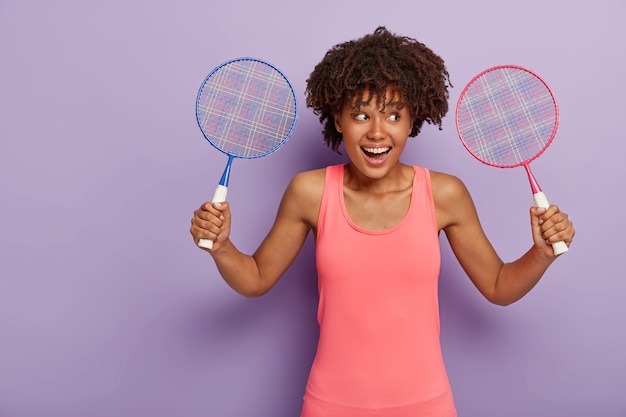 格好良い縮れ毛の若い女性は2つのテニスラケットを持って、ピンクのベストを着て、友達と好きなゲームをしたい、前向きに笑う