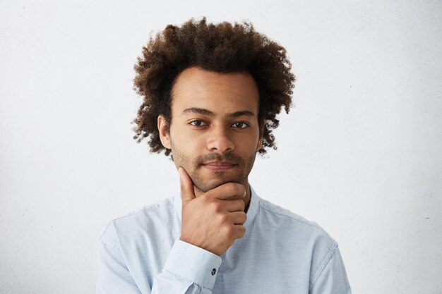 Good-looking dark-skinned male with curly hair having pensive expression holding hand on chin