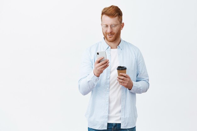 Good-looking european redhead man with bristle in glasses and shirt checking time in smartphone while standing over gray wall waiting for friend, drinking coffee, looking at cellphone screen, smiling