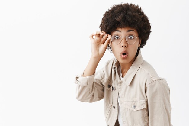 Good-looking female african american geek in glasses and beige shirt, touching rim of eyewear, folding lips and staring amazed, interested over gray wall