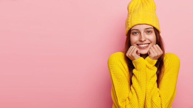 格好良い女性モデルは、両手であごを持ち、カメラに優しく微笑み、スタイリッシュな黄色の帽子とセーターを着て、ピンクの壁にモデルを置きます。