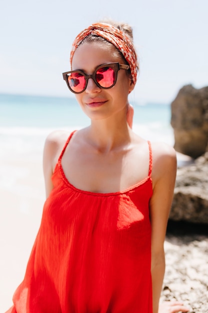 바다에 빨간 드레스를 입고 포즈를 취하는 잘 생긴 여성 모델. 야생 해변에서 아침을 보내는 트렌디 한 스파클 선글라스에 가볍게 무두질 한 젊은 아가씨.