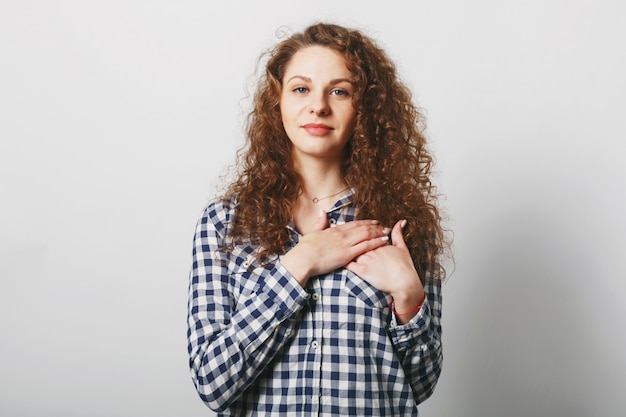 Good looking female model with curly bushy hair keeps hand on chest